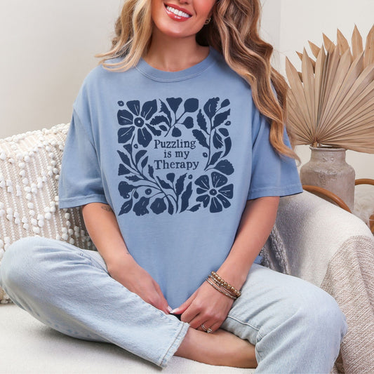 Model is wearing a Comfort Colors 1717 muted denim blue (washed denim) T-shirt. The shirt has a floral dark blue design that surrounds the words "Puzzling is my therapy"