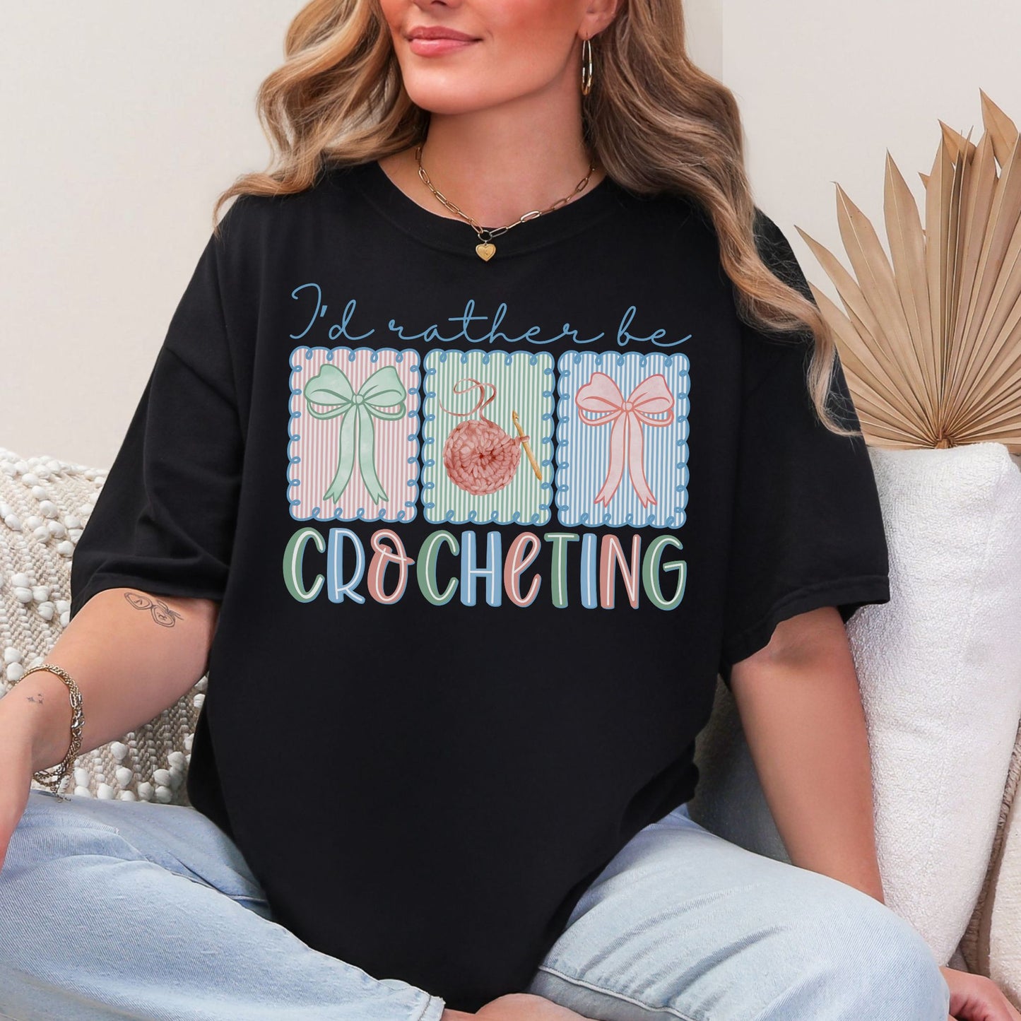 Model is wearing a Comfort Colors 1717 Black T-shirt featuring the phrase “I’d rather be crocheting” in soft pastel lettering, with three decorative panels above showing bows and a yarn ball with a crochet hook in light pink, mint green, and soft blue tones.