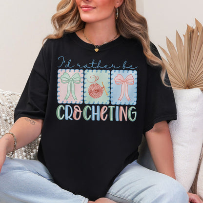Model is wearing a Comfort Colors 1717 Black T-shirt featuring the phrase “I’d rather be crocheting” in soft pastel lettering, with three decorative panels above showing bows and a yarn ball with a crochet hook in light pink, mint green, and soft blue tones.