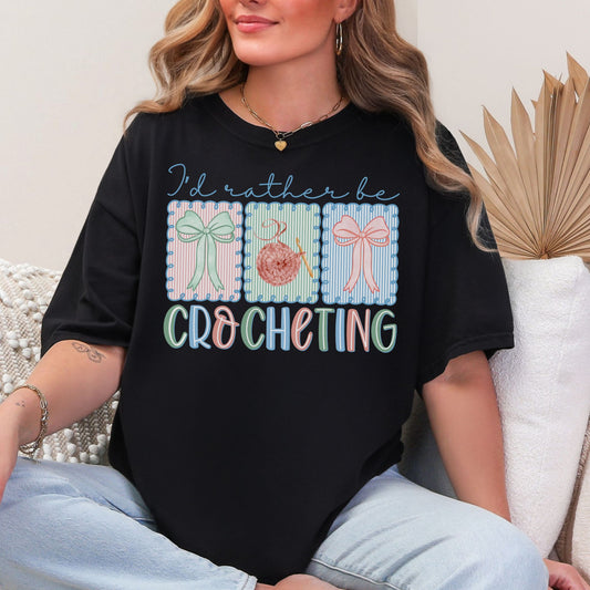 Model is wearing a Comfort Colors 1717 Black T-shirt featuring the phrase “I’d rather be crocheting” in soft pastel lettering, with three decorative panels above showing bows and a yarn ball with a crochet hook in light pink, mint green, and soft blue tones.