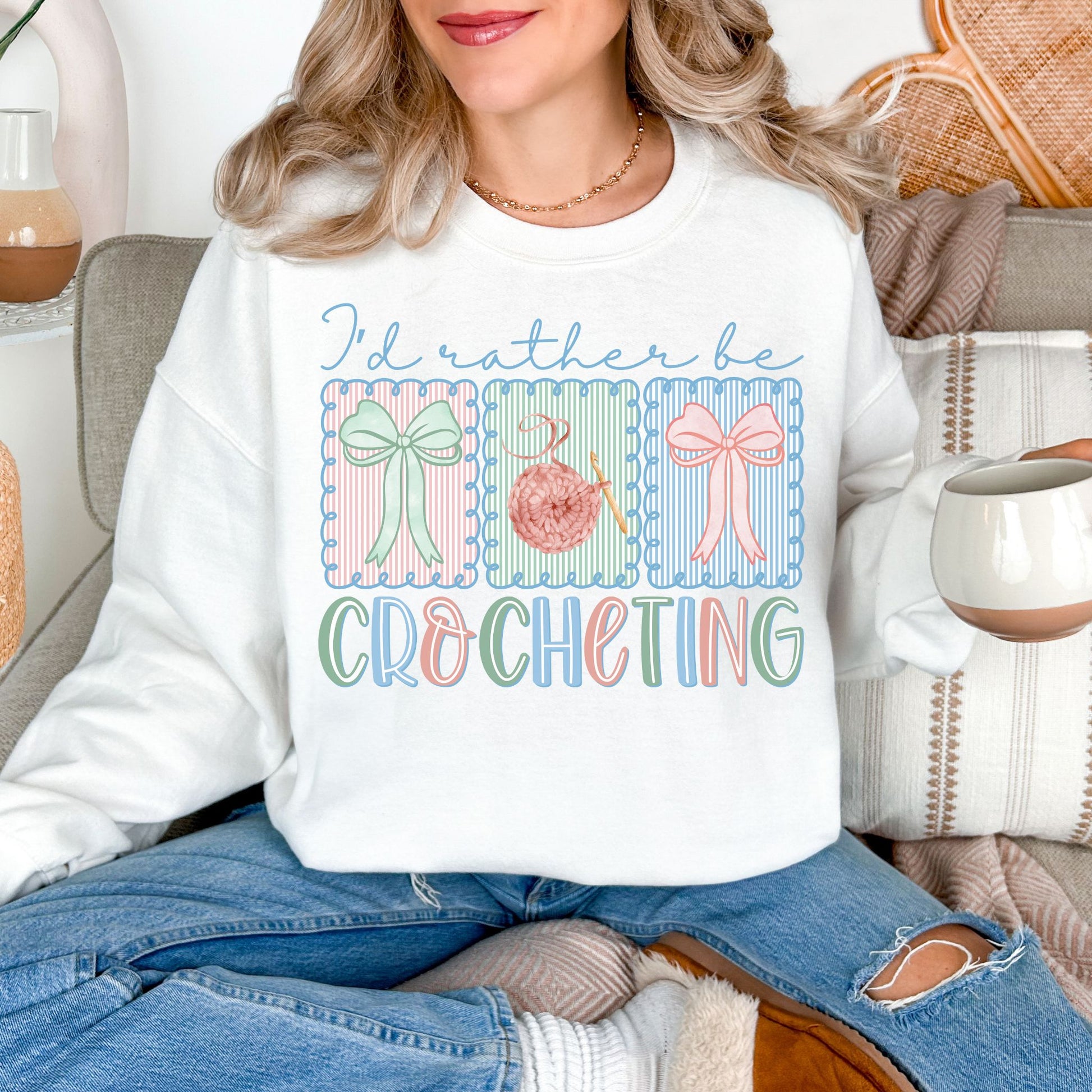 Model is wearing a Gildan 18000 bright white (white) Sweatshirt featuring the phrase “I’d rather be crocheting” in soft pastel lettering, with three decorative panels above showing bows and a yarn ball with a crochet hook in light pink, mint green, and soft blue tones.