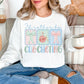 Model is wearing a Gildan 18000 bright white (white) Sweatshirt featuring the phrase “I’d rather be crocheting” in soft pastel lettering, with three decorative panels above showing bows and a yarn ball with a crochet hook in light pink, mint green, and soft blue tones.