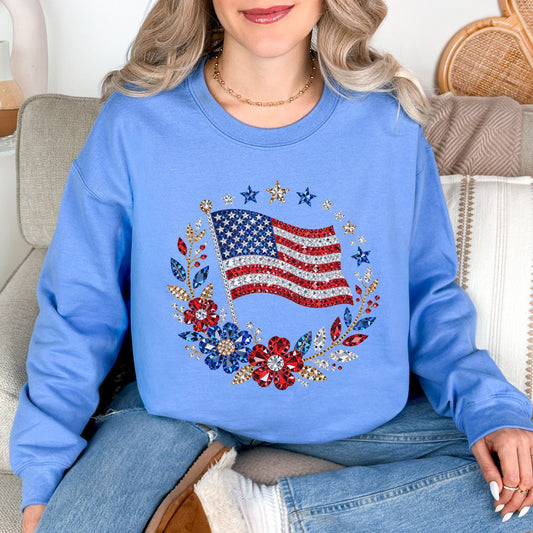 Model wearing a Gildan 18000 sweatshirt in light sky blue (carolina blue) featuring a faux rhinestone American flag design with diamond art-inspired gem details, surrounded by floral elements and star accents.