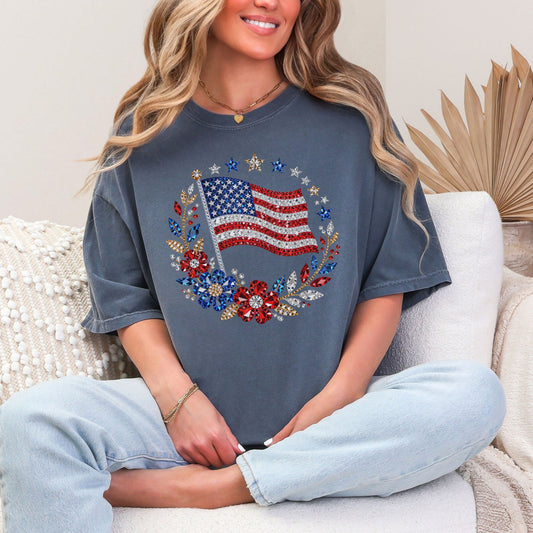 Model wearing a Comfort Colors 1717 t-shirt in muted medium blue (denim) featuring a faux rhinestone American flag design with diamond art-inspired gem details, surrounded by floral elements and star accents.