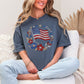 Model wearing a Comfort Colors 1717 t-shirt in muted medium blue (denim) featuring a faux rhinestone American flag design with diamond art-inspired gem details, surrounded by floral elements and star accents.