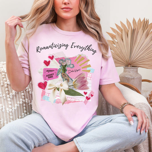Model is wearing a Comfort Colors 1717 T-Shirt in soft pink (blossom) featuring a Scrapbook-style collage graphic with soft pink (blush pink) paper, red (petal pink) hearts, a cream (soft beige) bow, and a pink (soft peach) tulip. Handwritten text reads “Romanticizing Everything” and “Date Night!” in a cozy junk journaling aesthetic.