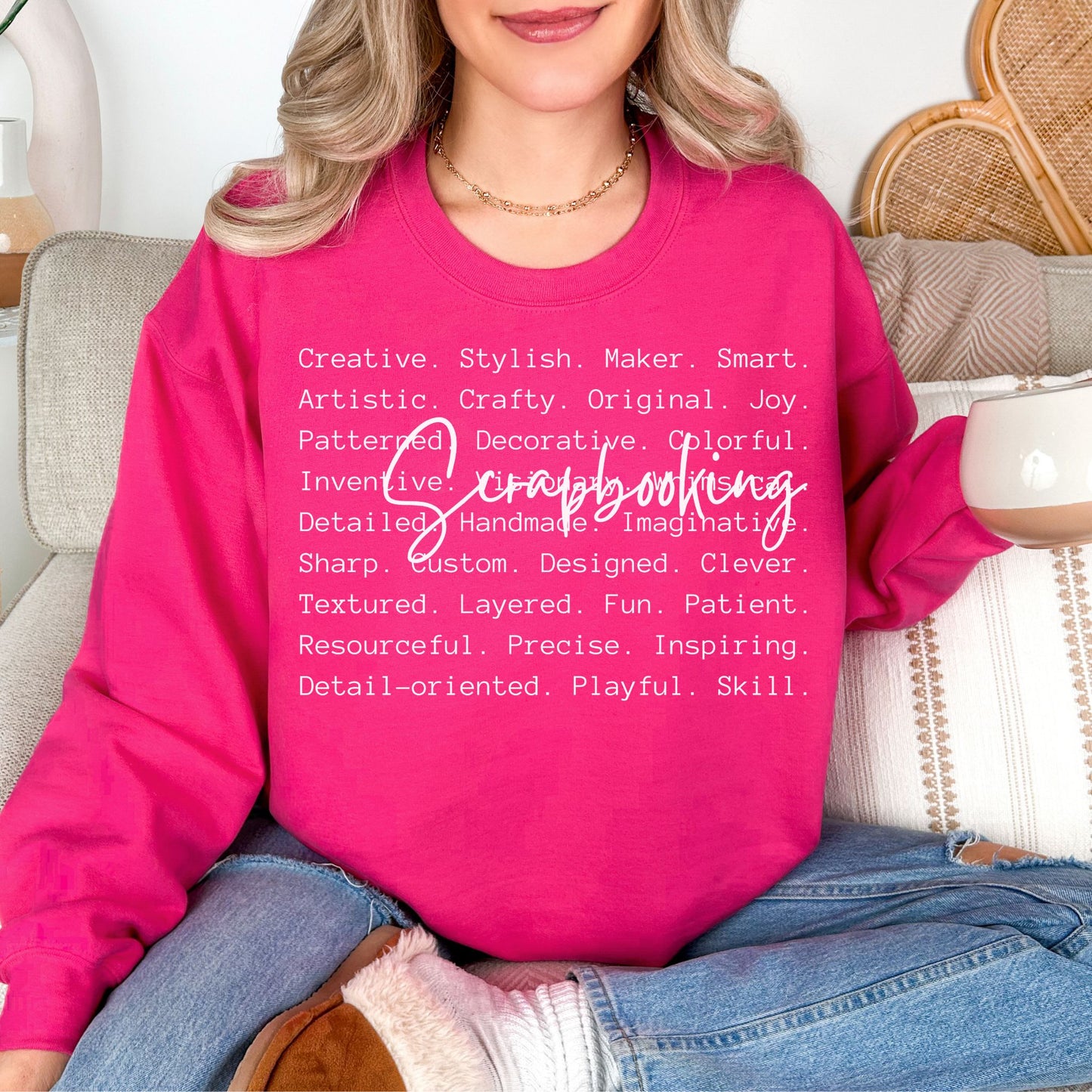 Model wearing a Gildan 18000 Sweatshirt in a bright pink (heliconia) color featuring a graphic design listing words that describe scrapbooking like "patient", "original", "joy" and "detailed". Over top of the words is the word "Scrapbooking" in bold cursive.