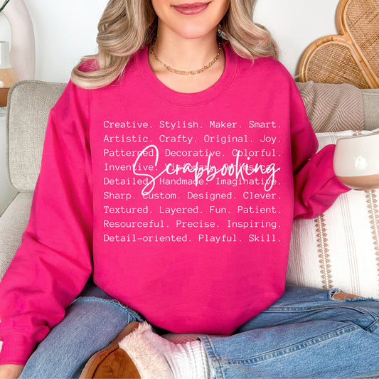 Model wearing a Gildan 18000 Sweatshirt in a bright pink (heliconia) color featuring a graphic design listing words that describe scrapbooking like "patient", "original", "joy" and "detailed". Over top of the words is the word "Scrapbooking" in bold cursive.