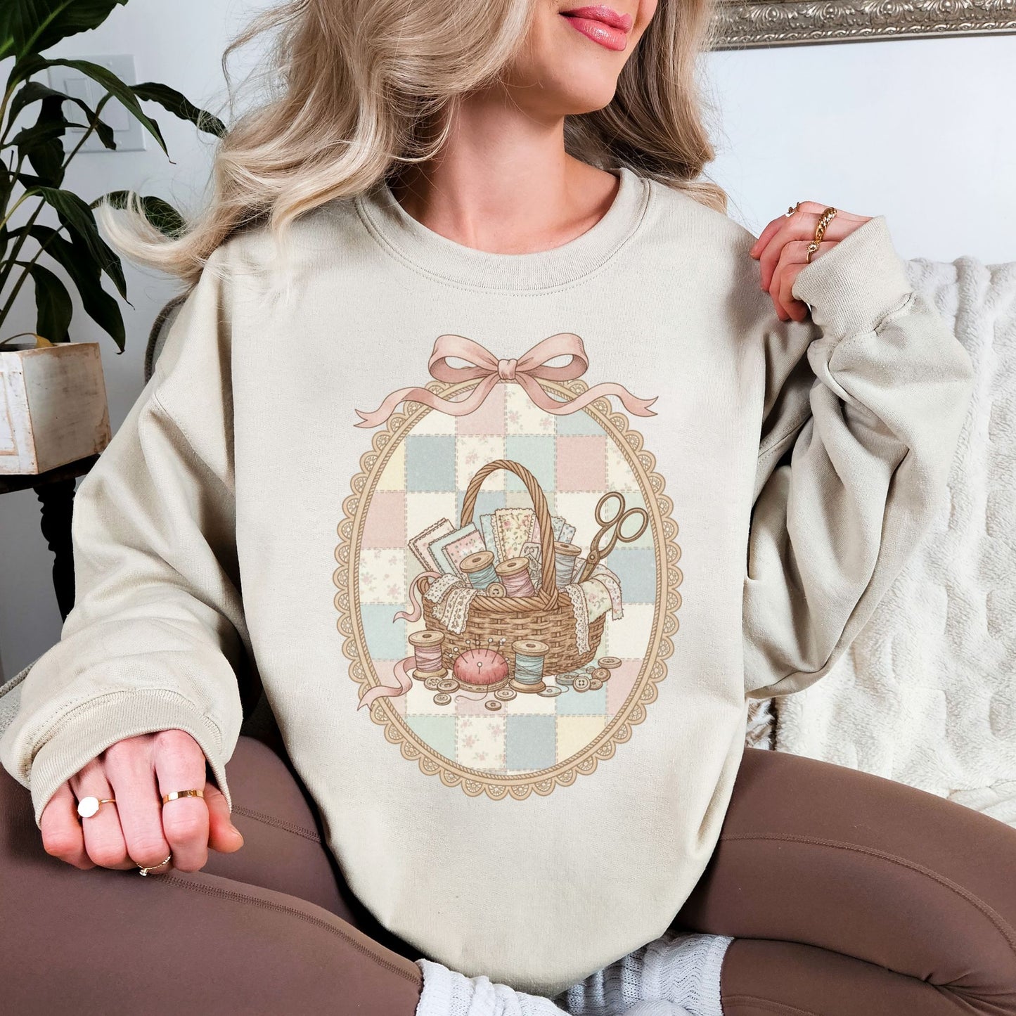 Model wearing a Gildan 18000 sweatshirt in light beige (sand) featuring a vintage coquette sewing design with a wicker basket filled with fabric, lace, thread spools, buttons, scissors, and sewing notions. The design is framed in an ornate oval with a soft pastel patchwork background and topped with a pink bow, giving a feminine shabby chic sewing aesthetic.