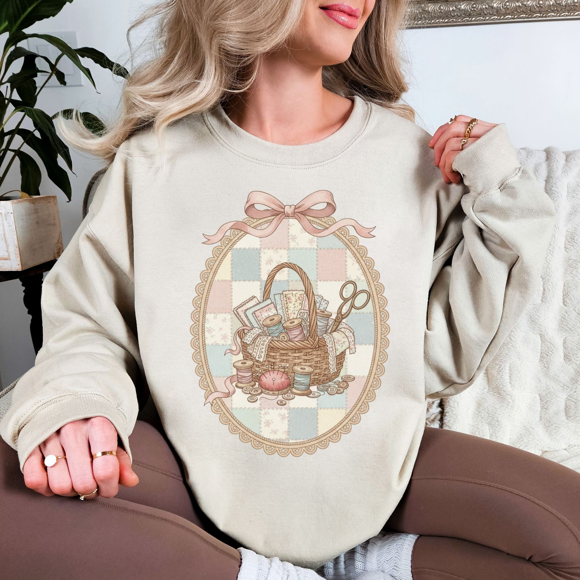 Model wearing a Gildan 18000 sweatshirt in light beige (sand) featuring a vintage coquette sewing design with a wicker basket filled with fabric, lace, thread spools, buttons, scissors, and sewing notions. The design is framed in an ornate oval with a soft pastel patchwork background and topped with a pink bow, giving a feminine shabby chic sewing aesthetic.