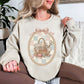 Model wearing a Gildan 18000 sweatshirt in light beige (sand) featuring a vintage coquette sewing design with a wicker basket filled with fabric, lace, thread spools, buttons, scissors, and sewing notions. The design is framed in an ornate oval with a soft pastel patchwork background and topped with a pink bow, giving a feminine shabby chic sewing aesthetic.