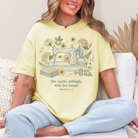 Model wearing a Comfort Colors 1717 in a soft pastel yellow (banana) featuring a graphic design of a vintage sewing machine and sewing supplies. In the background are faux pressed flowers and leaves. Below the graphic is text with the scripture "She works willingly with her hands Proverbs 31:13"