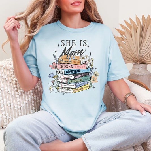 Model wearing a Comfort Colors 1717 soft medium blue (chambray) T-shirt. The shirt features a graphic design featuring the phrase “She is Mom” above a stack of colorful books labeled with affirmations including strong, chosen, beautiful, capable, victorious, never alone, and greatly loved, surrounded by flowers and sparkles in a faith-inspired style.