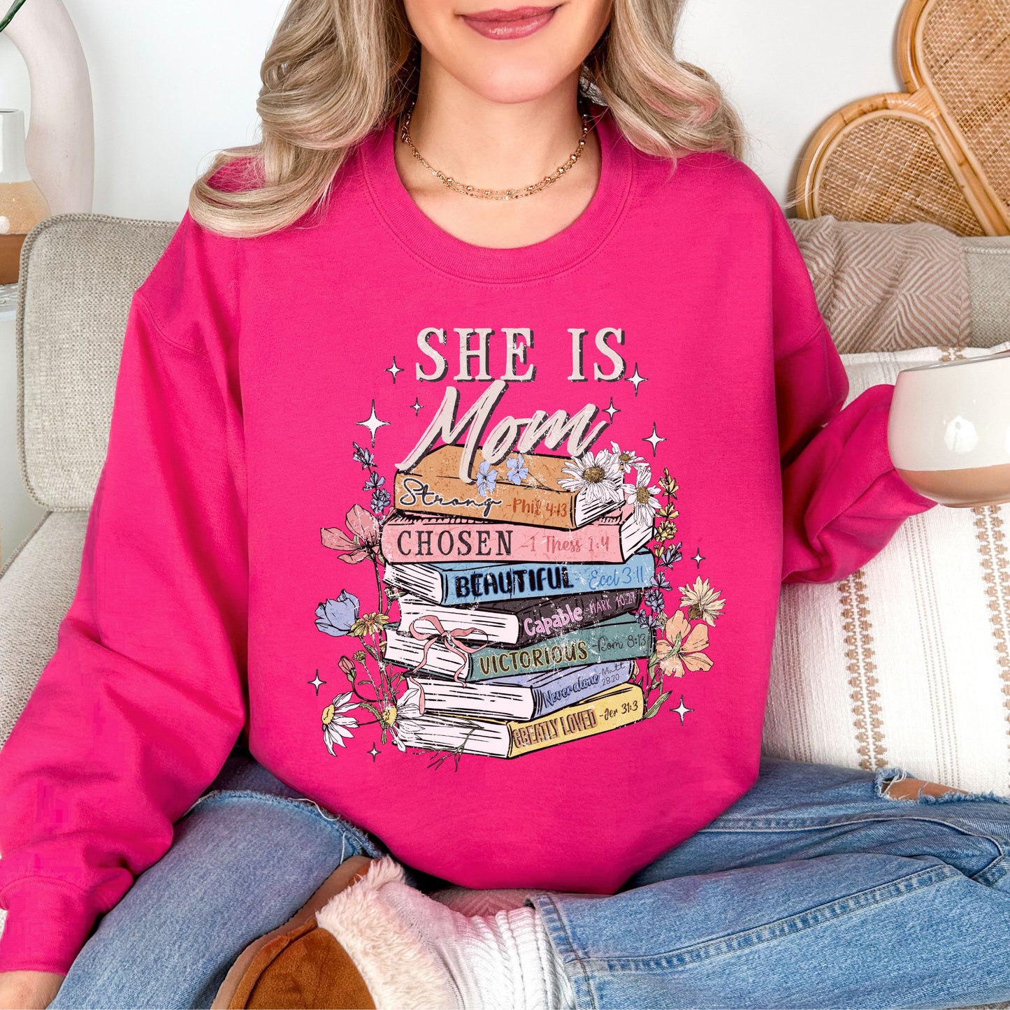 Model wearing a Gildan 18000 bright pink (heliconia) Sweatshirt. The sweatshirt features a graphic design featuring the phrase “She is Mom” above a stack of colorful books labeled with affirmations including strong, chosen, beautiful, capable, victorious, never alone, and greatly loved, surrounded by flowers and sparkles in a faith-inspired style.