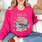 Model wearing a Gildan 18000 bright pink (heliconia) Sweatshirt. The sweatshirt features a graphic design featuring the phrase “She is Mom” above a stack of colorful books labeled with affirmations including strong, chosen, beautiful, capable, victorious, never alone, and greatly loved, surrounded by flowers and sparkles in a faith-inspired style.