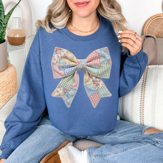 Model is wearing a Comfort Colors 1717 in rich medium-dark blue (indigo blue) sweatshirt featuring a faux patchwork-style bow made of pastel fabric-inspired patterns including florals, gingham, stripes, and polka dots in pink, blue, green, and yellow tones. The design is printed to resemble quilted patchwork and stitched fabric but is not real fabric or textured appliqué.