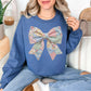 Model is wearing a Comfort Colors 1717 in rich medium-dark blue (indigo blue) sweatshirt featuring a faux patchwork-style bow made of pastel fabric-inspired patterns including florals, gingham, stripes, and polka dots in pink, blue, green, and yellow tones. The design is printed to resemble quilted patchwork and stitched fabric but is not real fabric or textured appliqué.