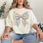Model is wearing a Comfort Colors 1717 in soft cream (ivory) T-shirt featuring a faux patchwork-style bow made of pastel fabric-inspired patterns including florals, gingham, stripes, and polka dots in pink, blue, green, and yellow tones. The design is printed to resemble quilted patchwork and stitched fabric but is not real fabric or textured appliqué.