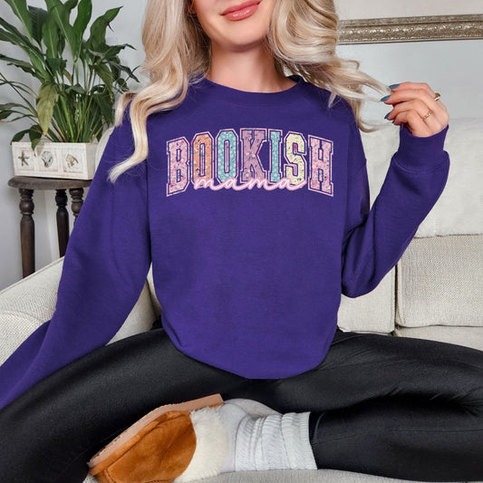 Model is wearing a Gildan 18000 sweatshirt in medium purple (purple). The shirt features a graphic design that says, "Bookish mama". "Bookish" are in a varsity letter font filled in with a beautiful pastel spring floral design.