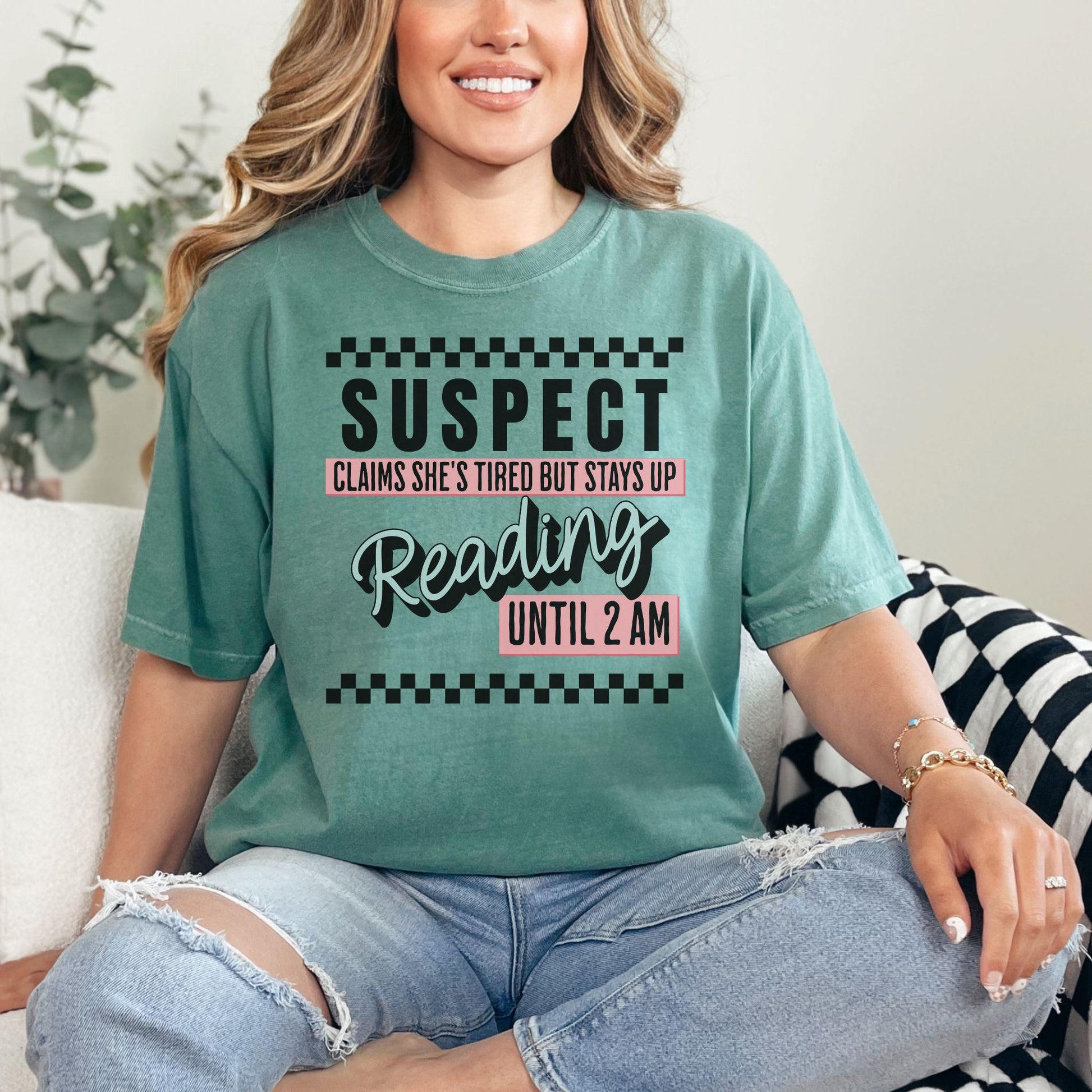 Model wearing a soft light green (light green) Comfort Colors t-shirt featuring wording that says "Suspect claims she's tired but stays up reading until 2 am" printed graphic with checkered strips on the top and bottom of the graphic.