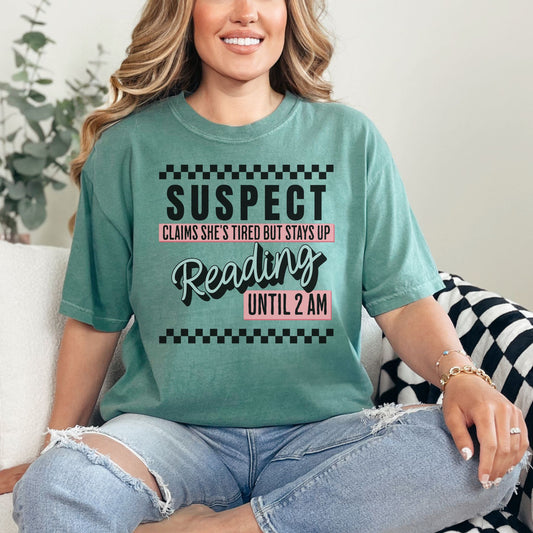 Model wearing a soft light green (light green) Comfort Colors t-shirt featuring wording that says "Suspect claims she's tired but stays up reading until 2 am" printed graphic with checkered strips on the top and bottom of the graphic.