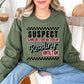 Model wearing a muted olive green (military green) Gildan Sweatshirt featuring wording that says "Suspect claims she's tired but stays up reading until 2 am" printed graphic with checkered strips on the top and bottom of the graphic.