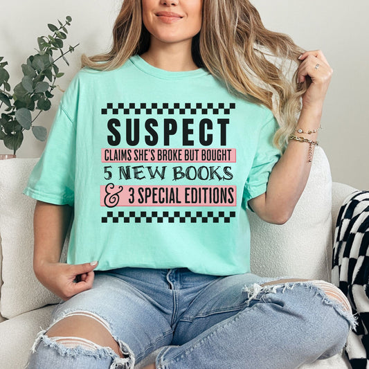 Model wearing a soft aqua blue (island reef) Comfort Colors t-shirt featuring wording that says "Suspect claims she's tired but stays up reading until 2 am" printed graphic with checkered strips on the top and bottom of the graphic.