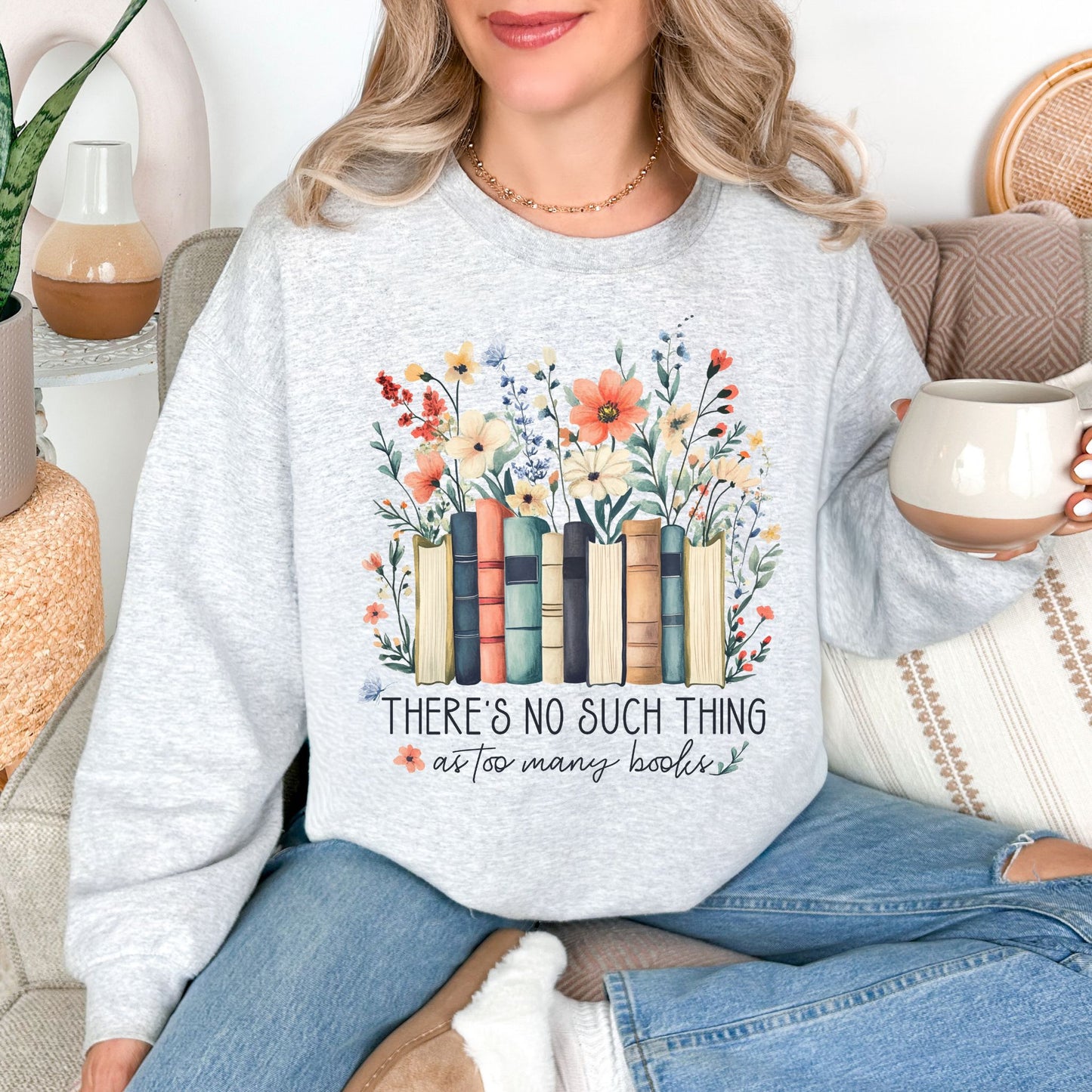 Model wearing a Gildan 18000 sweatshirt in very light gray (ash) featuring books lined up with wildflower florals behind it.  Underneath is the phrase "There's no such thing as too many books" in a mix of script and cursive fonts. 