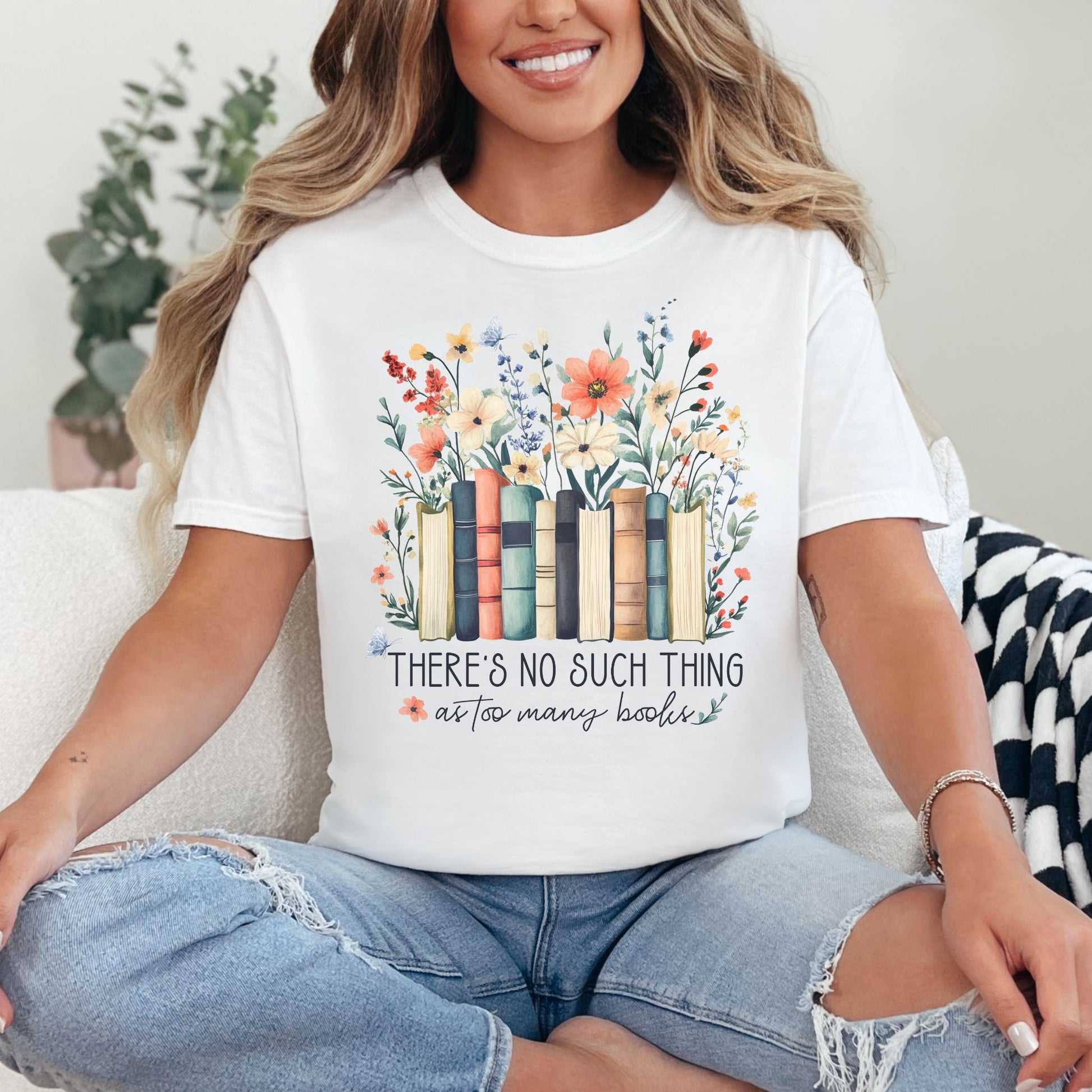 Model wearing a Comfort Colors 1717 in bright white (white) featuring books lined up with wildflower florals behind it.  Underneath is the phrase "There's no such thing as too many books" in a mix of script and cursive fonts. 