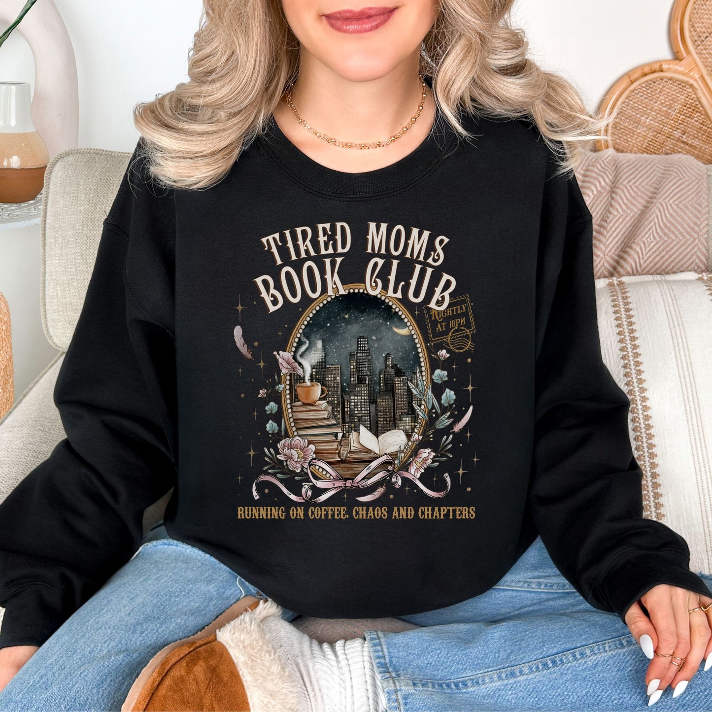 Model is wearing a deep black (Black) Gildan 18000 sweatshirt with a cozy bookish illustration featuring a nighttime city skyline framed by stacked books, a steaming coffee cup, florals, and ribbon accents in warm gold, soft pink, and muted teal tones. The text reads “Tired Moms Book Club” with “Running on Coffee, Chaos and Chapters” below.