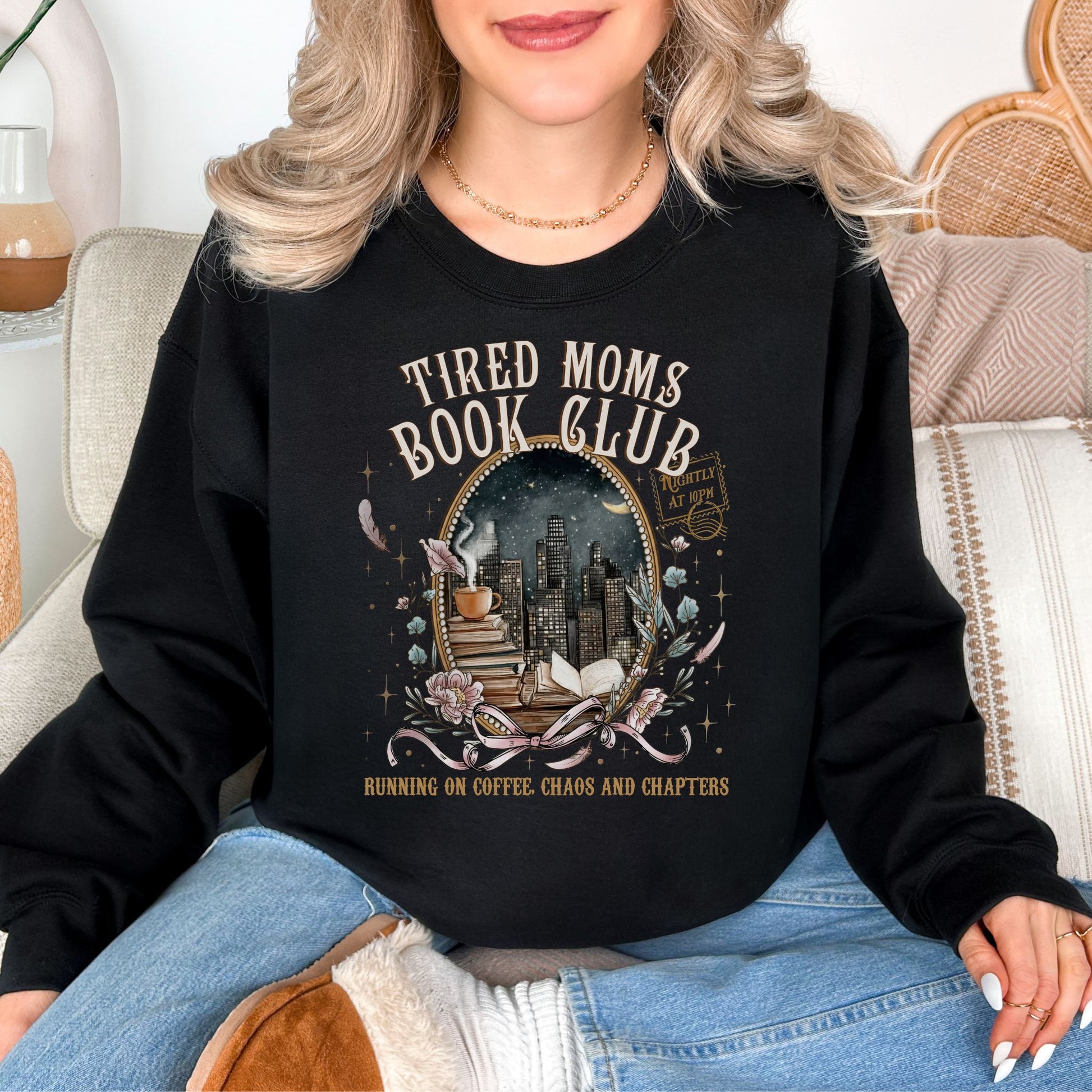 Model is wearing a deep black (Black) Gildan 18000 sweatshirt with a cozy bookish illustration featuring a nighttime city skyline framed by stacked books, a steaming coffee cup, florals, and ribbon accents in warm gold, soft pink, and muted teal tones. The text reads “Tired Moms Book Club” with “Running on Coffee, Chaos and Chapters” below.