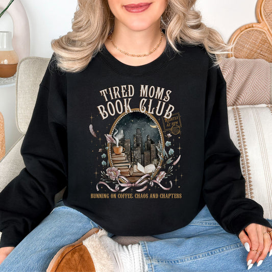 Model is wearing a deep black (Black) Gildan 18000 sweatshirt with a cozy bookish illustration featuring a nighttime city skyline framed by stacked books, a steaming coffee cup, florals, and ribbon accents in warm gold, soft pink, and muted teal tones. The text reads “Tired Moms Book Club” with “Running on Coffee, Chaos and Chapters” below.
