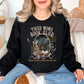 Model is wearing a deep black (Black) Gildan 18000 sweatshirt with a cozy bookish illustration featuring a nighttime city skyline framed by stacked books, a steaming coffee cup, florals, and ribbon accents in warm gold, soft pink, and muted teal tones. The text reads “Tired Moms Book Club” with “Running on Coffee, Chaos and Chapters” below.