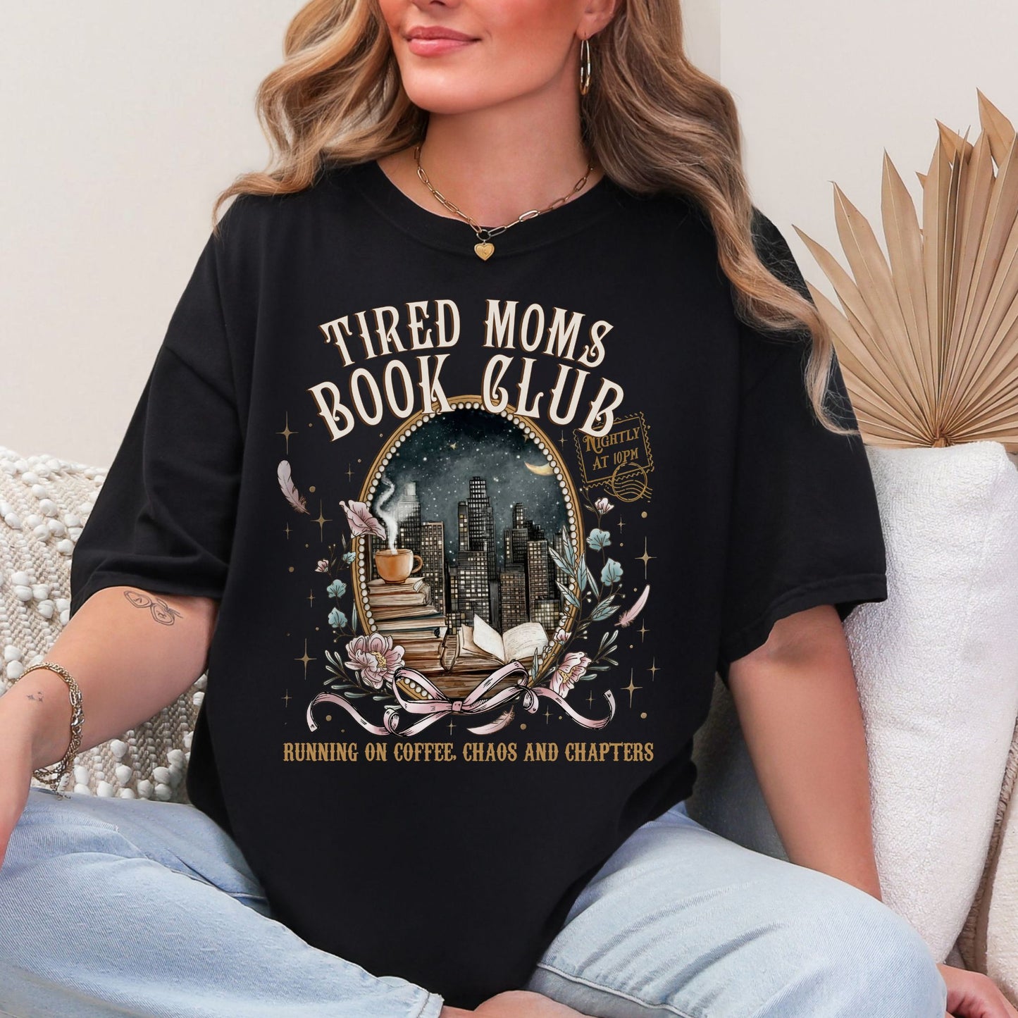 Model is wearing a Black Comfort Colors 1717 T-shirt with a cozy bookish illustration featuring a nighttime city skyline framed by stacked books, a steaming coffee cup, florals, and ribbon accents in warm gold, soft pink, and muted teal tones. The text reads “Tired Moms Book Club” with “Running on Coffee, Chaos and Chapters” below.