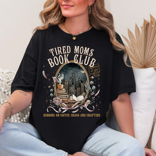 Model is wearing a Black Comfort Colors 1717 T-shirt with a cozy bookish illustration featuring a nighttime city skyline framed by stacked books, a steaming coffee cup, florals, and ribbon accents in warm gold, soft pink, and muted teal tones. The text reads “Tired Moms Book Club” with “Running on Coffee, Chaos and Chapters” below.