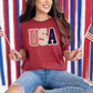 Model wearing a Comfort Colors1717 t-shirt in muted brick red (chili) with a patriotic “USA” design featuring striped, knit-style, and star patterns accented with lace-style borders in a faux embroidery style.