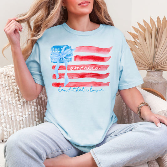 Model wearing a Comfort Colors 1717 in soft medium blue (chambray) featuring a watercolor American flag design with soft red and blue brushstroke stripes, a blue bow detail, and script text.