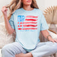 Model wearing a Comfort Colors 1717 in soft medium blue (chambray) featuring a watercolor American flag design with soft red and blue brushstroke stripes, a blue bow detail, and script text.