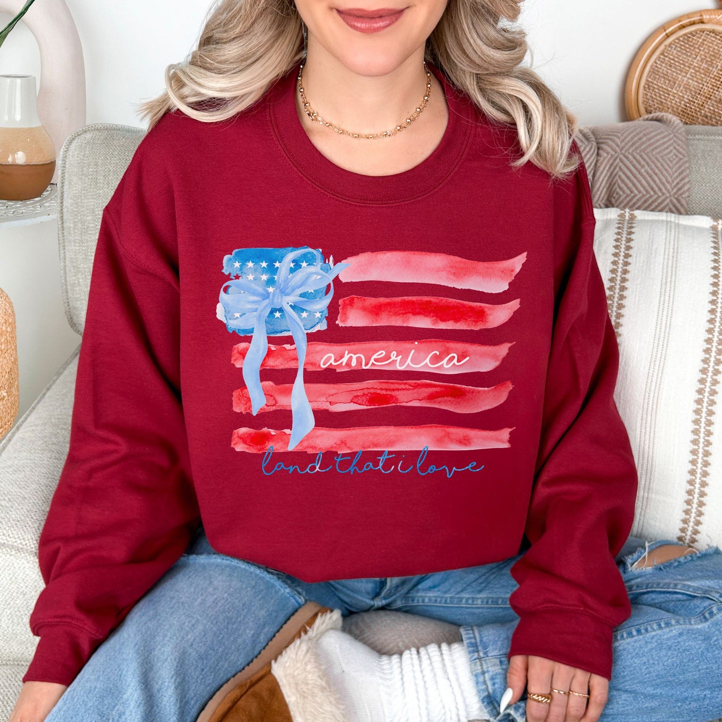 Model wearing a Gildan 18000 in dark red (cardinal red) featuring a watercolor American flag design with soft red and blue brushstroke stripes, a blue bow detail, and script text.
